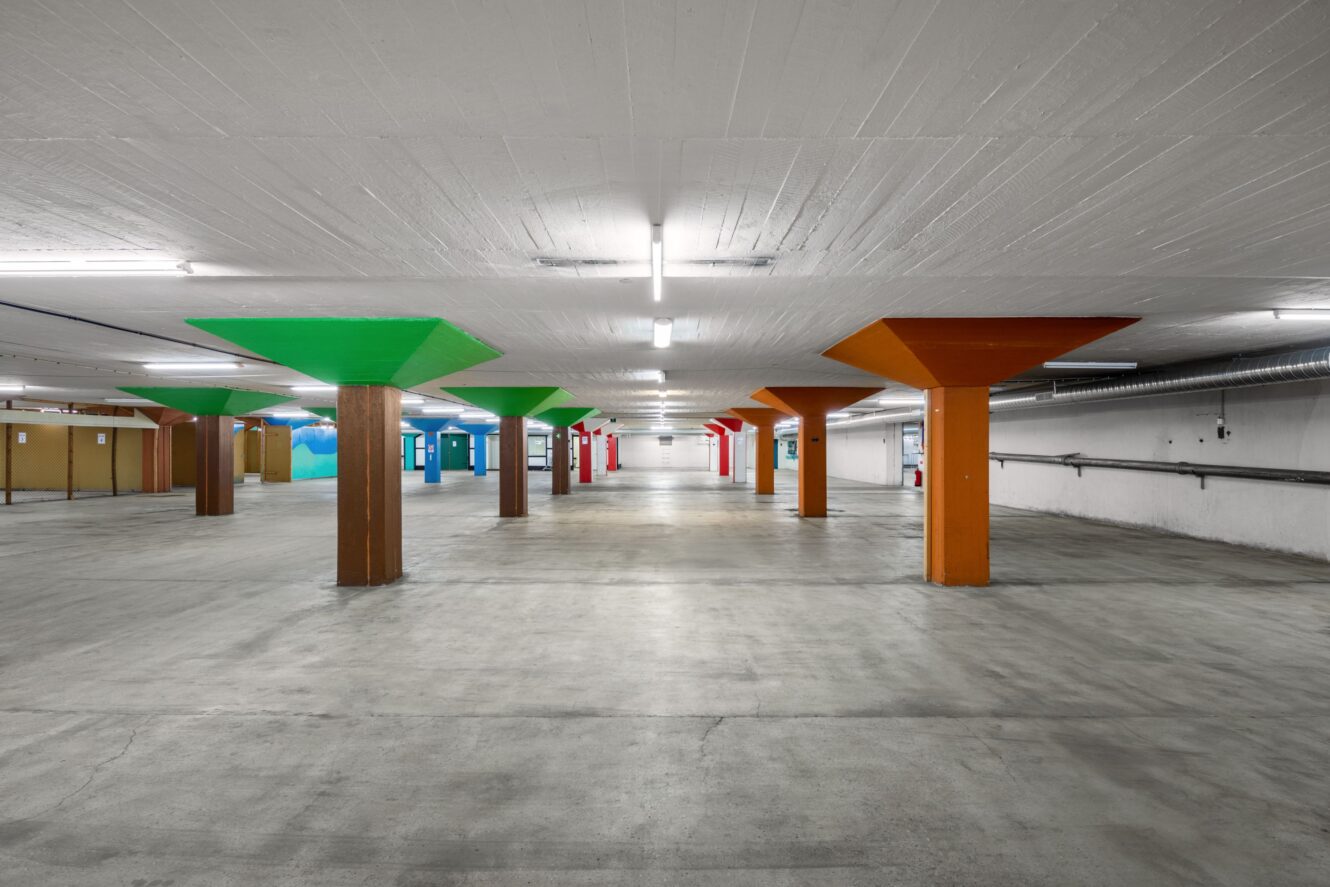Tomt parkeringsgarage i betong med fyrkantiga pelare målade i grönt, orange, rött och blått, under starka lysrör i taket.