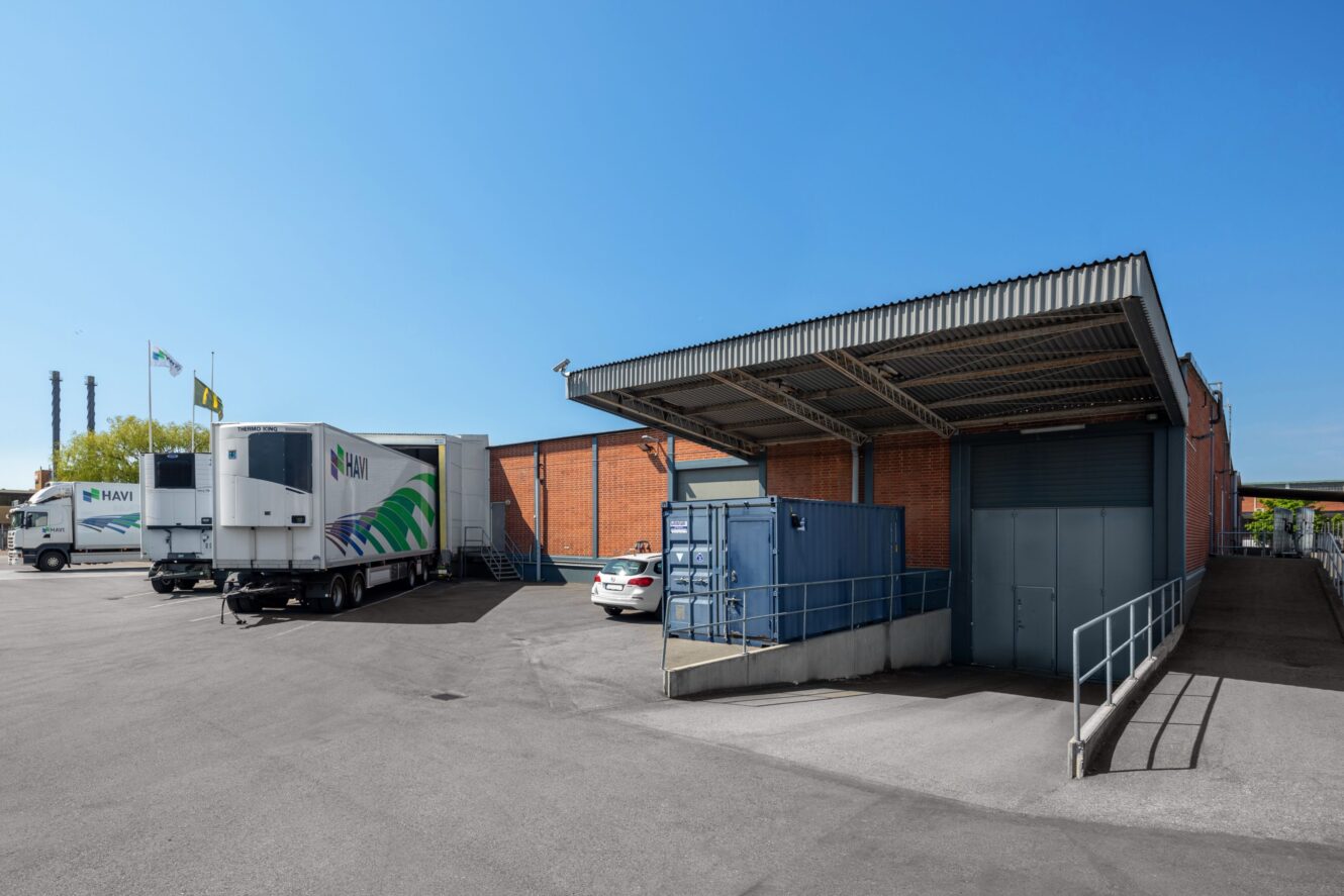 Ett lastkajsområde med tegelbyggnader, parkerade lastbilar, en vit bil, en blå fraktcontainer och en täckt ramp under en klarblå himmel.