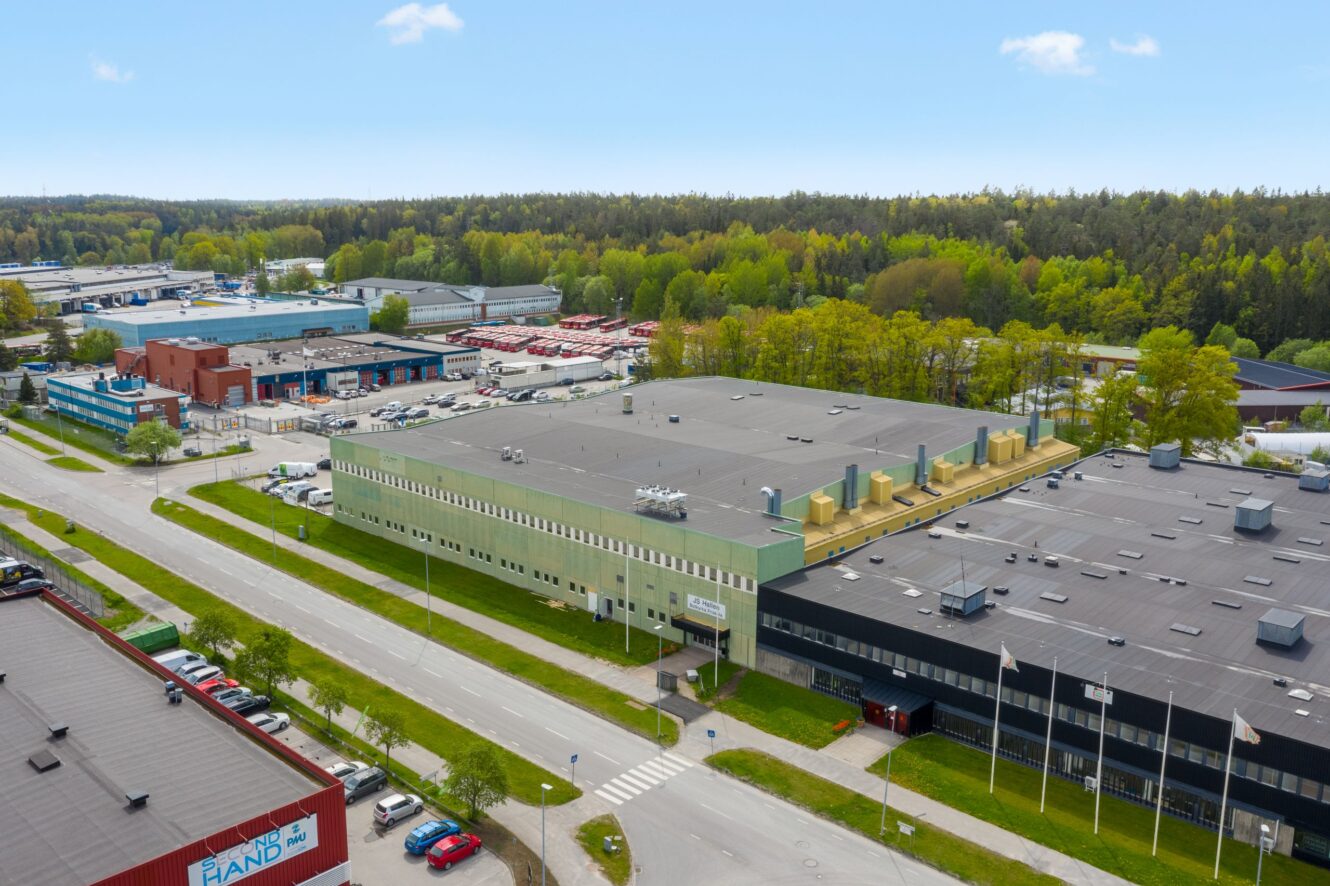 Flygbild över ett industriområde med stora lagerbyggnader, intilliggande vägar, parkerade fordon och omgivande träd under en klar himmel.