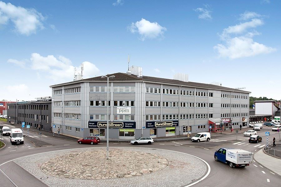 En stor grå kontorsbyggnad med flera fönster står nära en livligt trafikerad rondell, med flera bilar och skåpbilar synliga på vägen under en delvis molnig himmel.