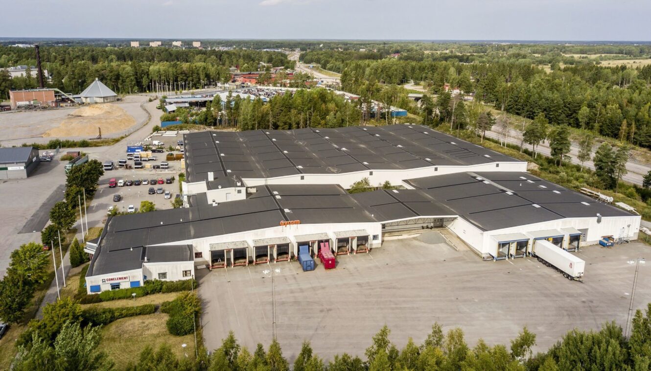 Flygfoto över ett stort industriellt lagerkomplex omgivet av träd, parkeringsplatser och flera lastbryggor med lastbilar och containrar.