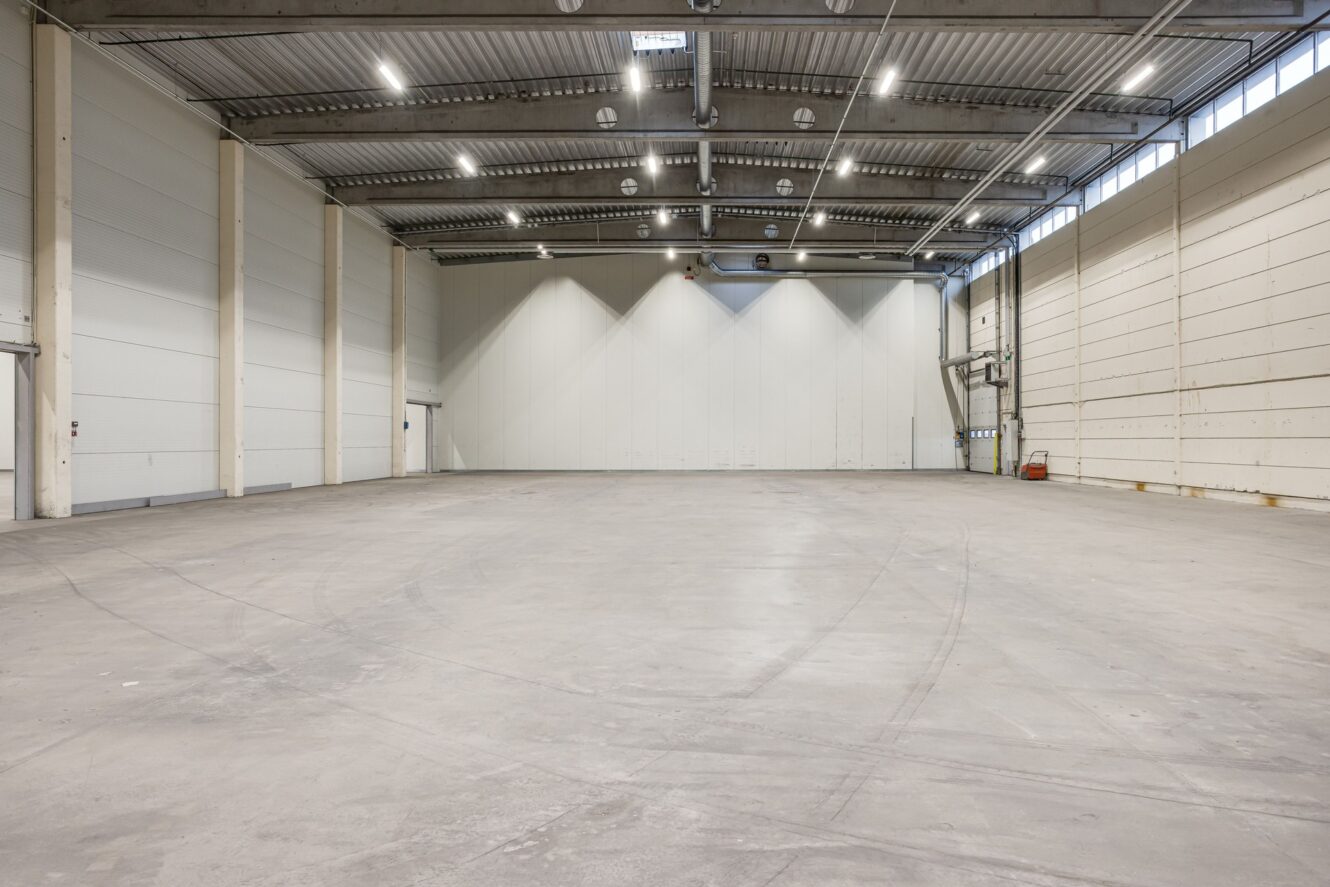 Stor tom lagerlokal med högt i tak, betonggolv och vita väggar, upplyst av taklampor och naturligt ljus från höga fönster på höger sida.