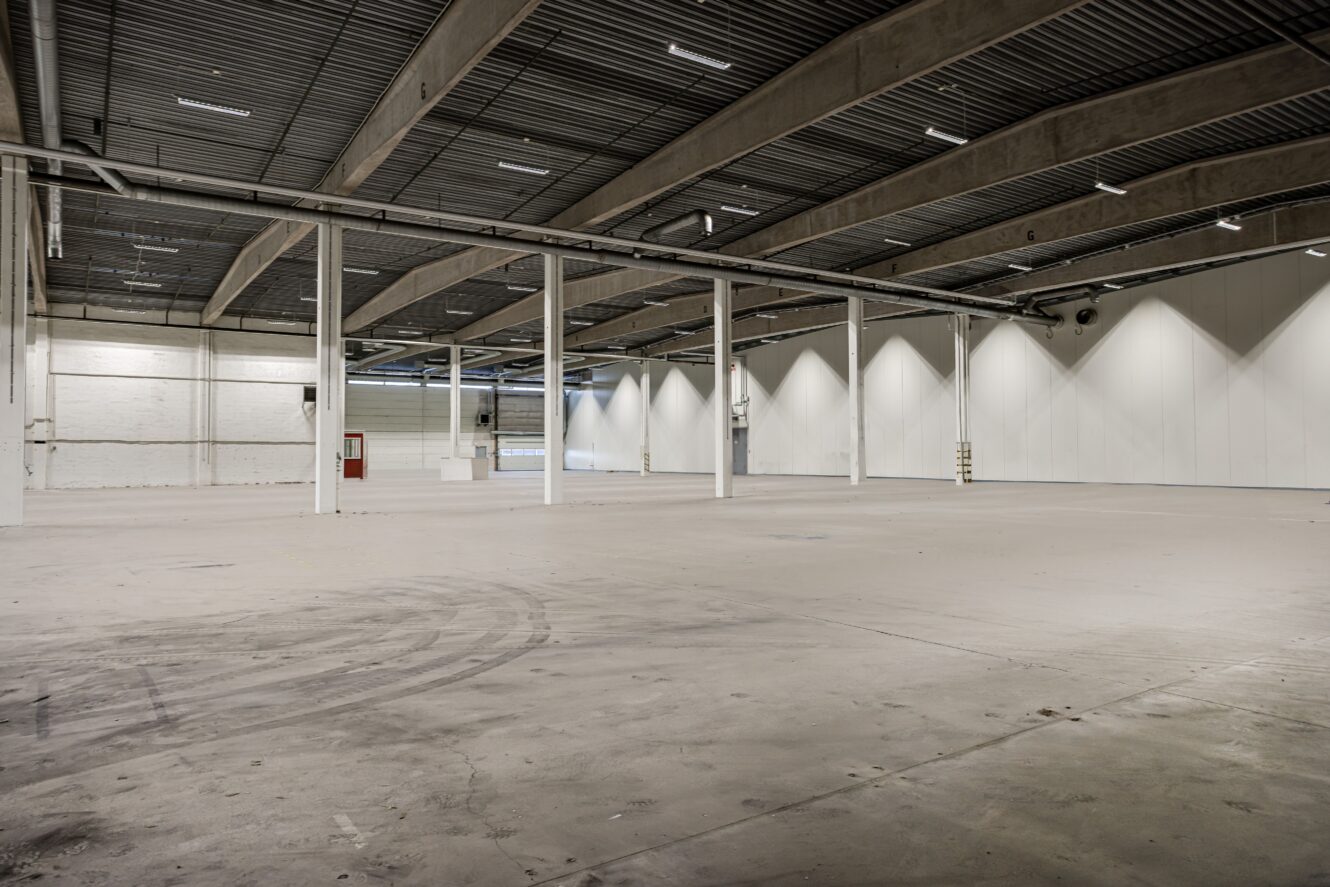 En stor, tom lagerlokal med betonggolv, högt i tak, stödpelare och vita väggar i fluorescerande belysning.