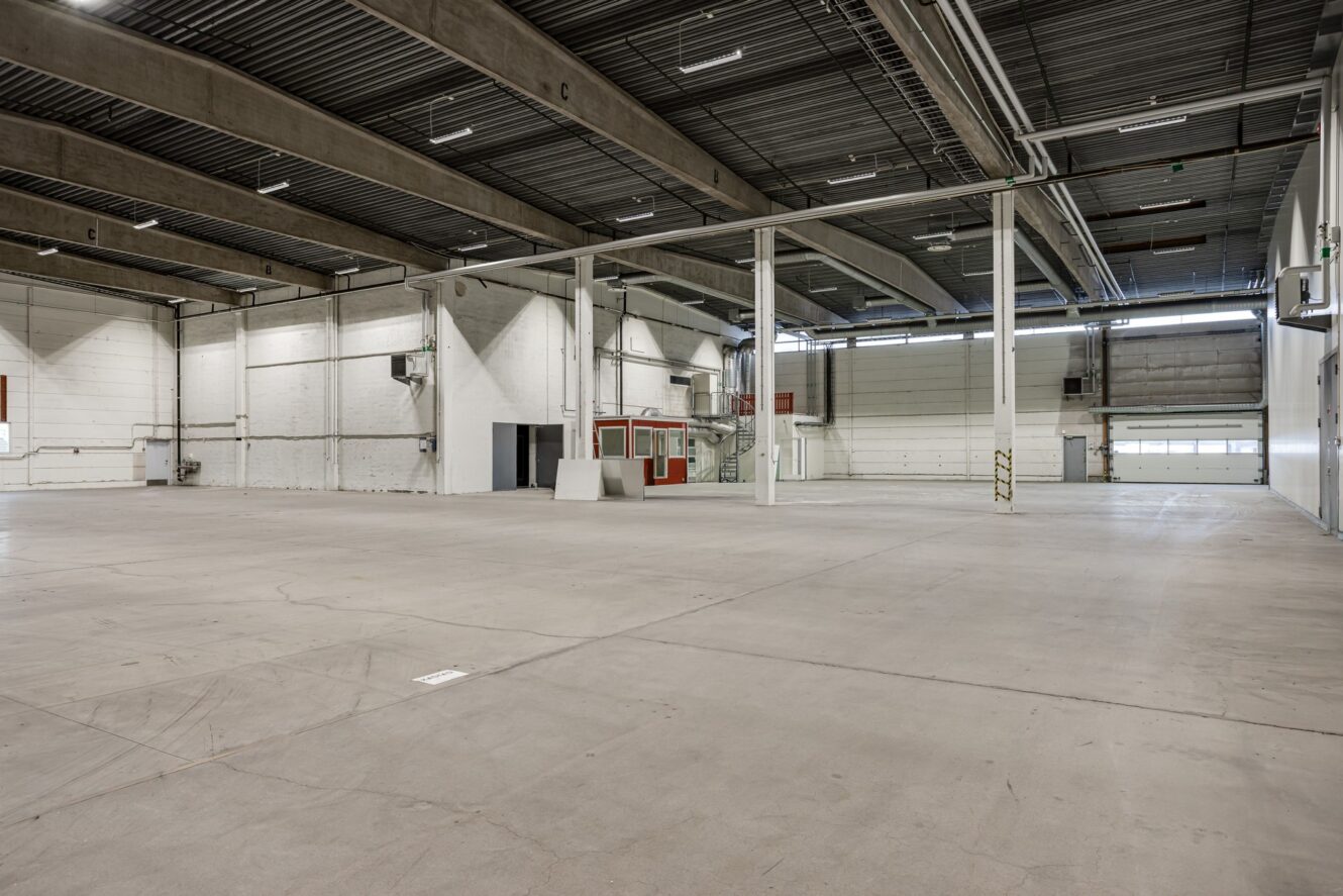 Stort, tomt industrilager med betonggolv, högt i tak, synliga bjälkar, takbelysning och en liten röd kontorsbyggnad nära den bakre väggen.