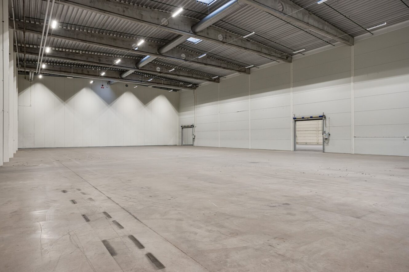 Rymlig tom lagerlokal med högt i tak, vita väggar, betonggolv och två stora dörrar till lastkajer.