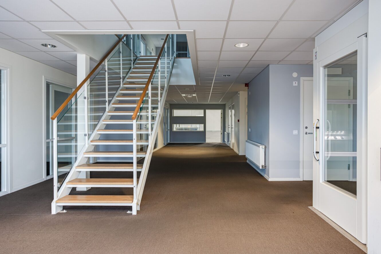 Interiör i en modern kontorsbyggnad med heltäckningsmattor, en trappa med glasräcke, vita väggar och stora fönster som ger naturligt ljus.