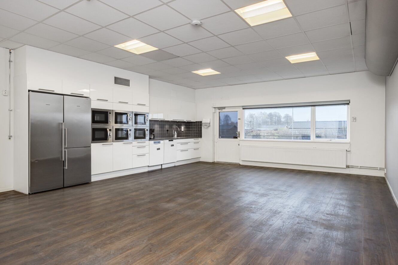 Rymligt, modernt kök med vita skåp, vitvaror i rostfritt stål, mörkt trägolv och stort fönster som släpper in naturligt ljus.