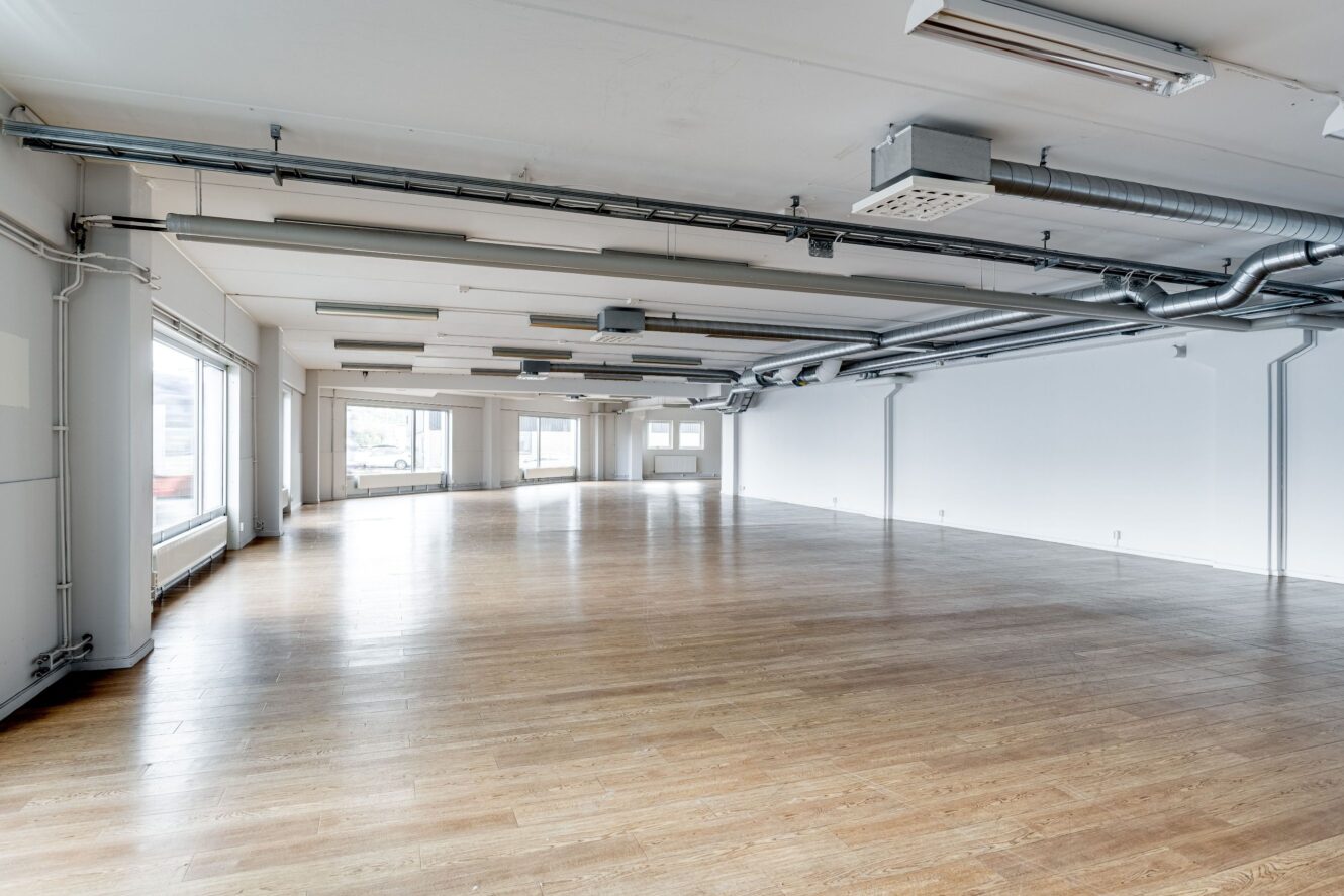 Rymligt tomt rum med trägolv, vita väggar, stora fönster och synliga takkanaler och belysningsarmaturer.