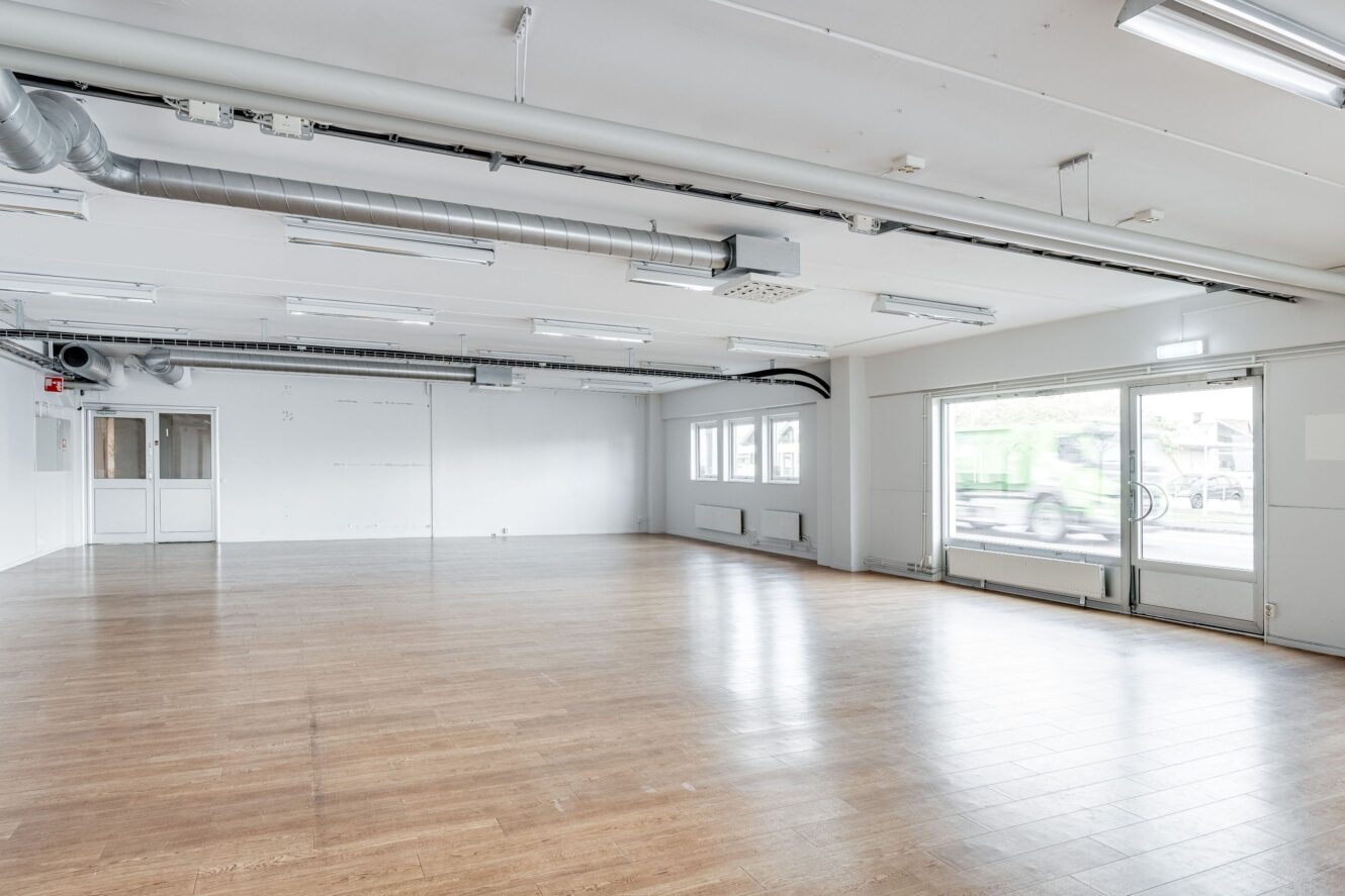 Ett rymligt, tomt rum med trägolv, vita väggar, stora fönster och synliga takkanaler.