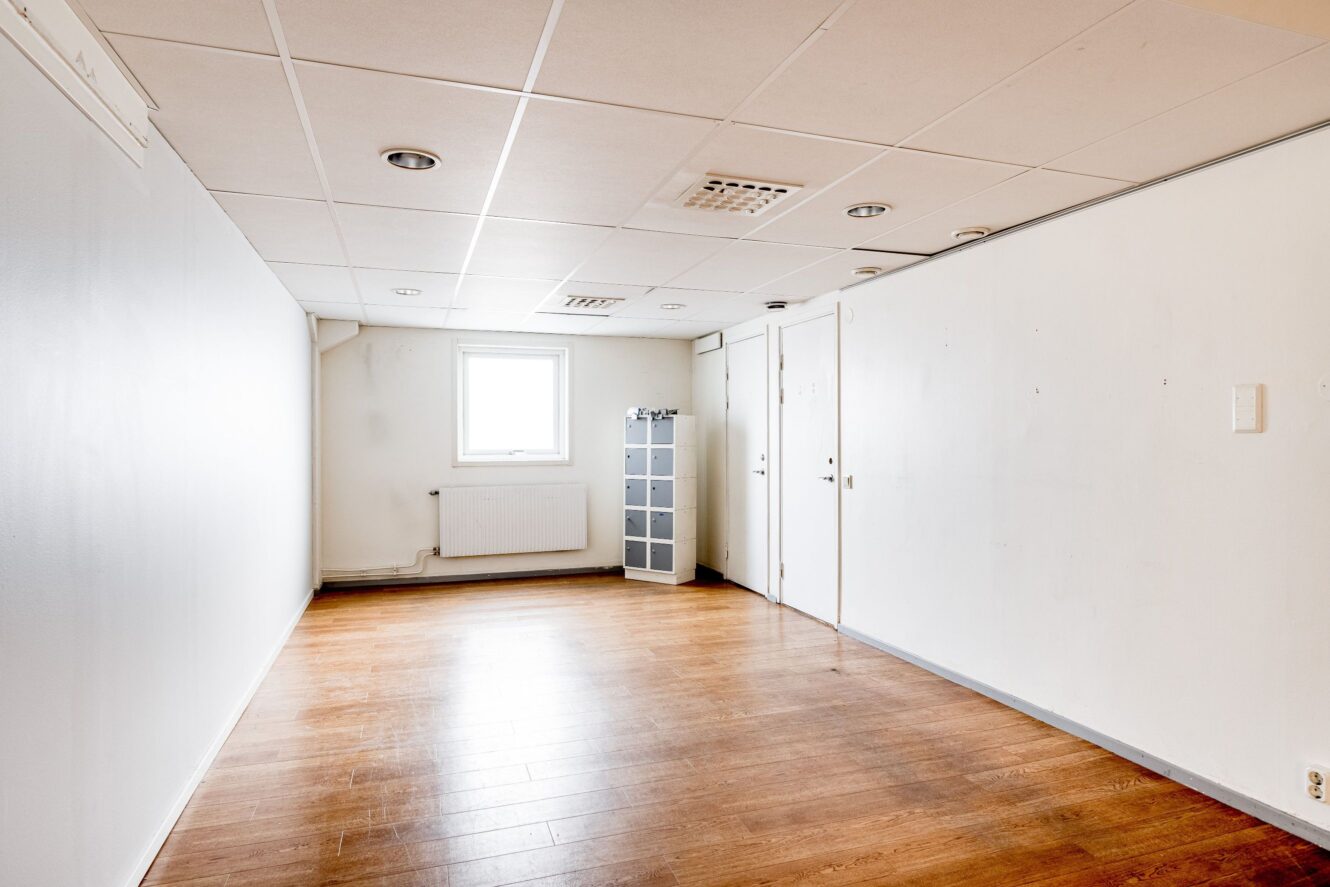 Tomt rum med vita väggar, ljust trägolv, ett enda fönster, taklampor och en liten grå hylla mot den bortre väggen.