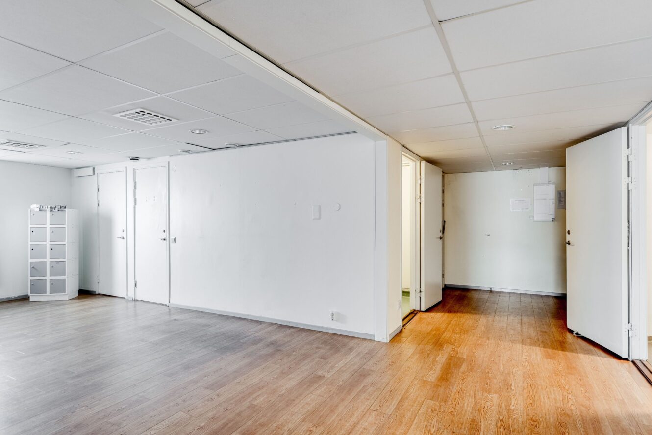 Tomt kontorsutrymme med vita väggar, trägolv, takpaneler, flera öppna dörrar och en liten uppsättning skåp i hörnet.