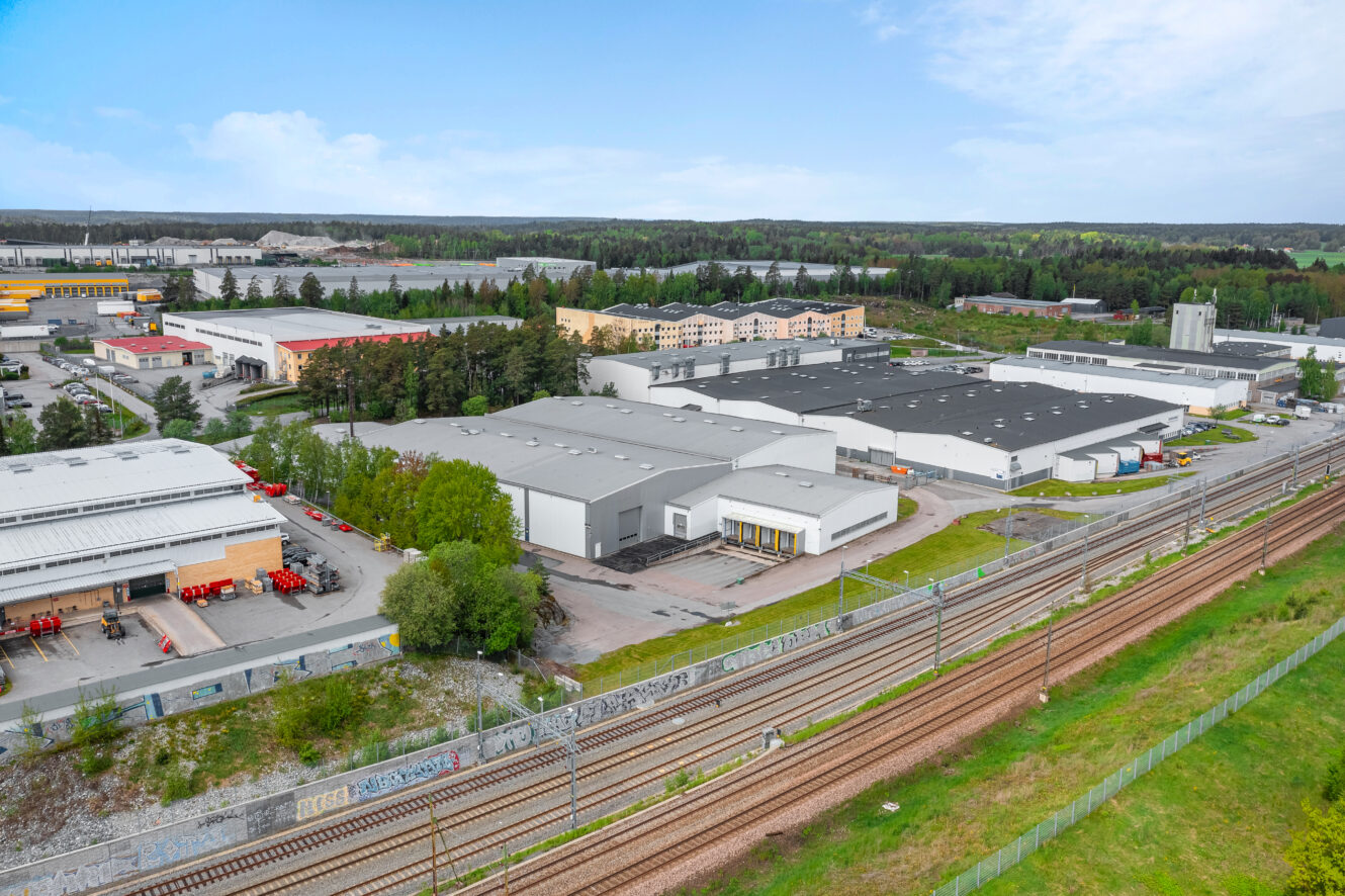 Flygfoto över ett industriområde med stora lagerlokaler, tågspår i förgrunden och skogsklädda omgivningar under en delvis molnig himmel.