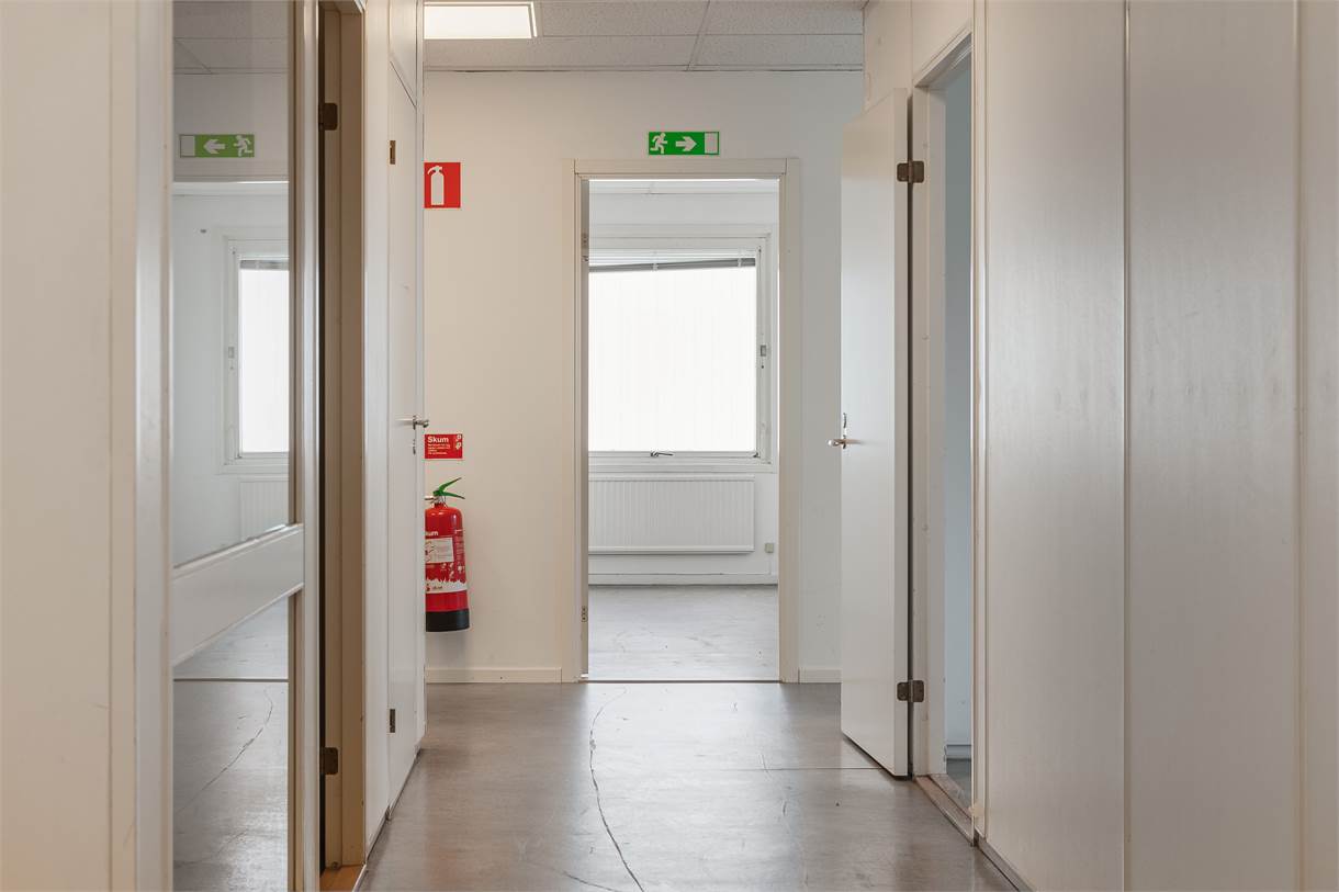 En korridor med öppna vita dörrar, en brandsläckare monterad på väggen och gröna nödutgångsskyltar ovanför dörröppningarna.