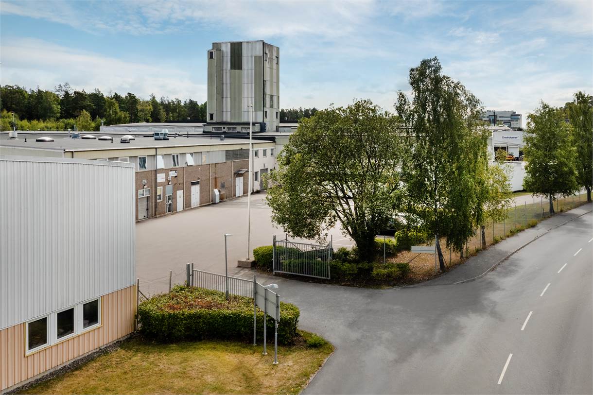 Ett industriområde med lagerlokaler, asfalterad tomt och en hög betongbyggnad i bakgrunden; träd och en väg löper längs med fastigheten.