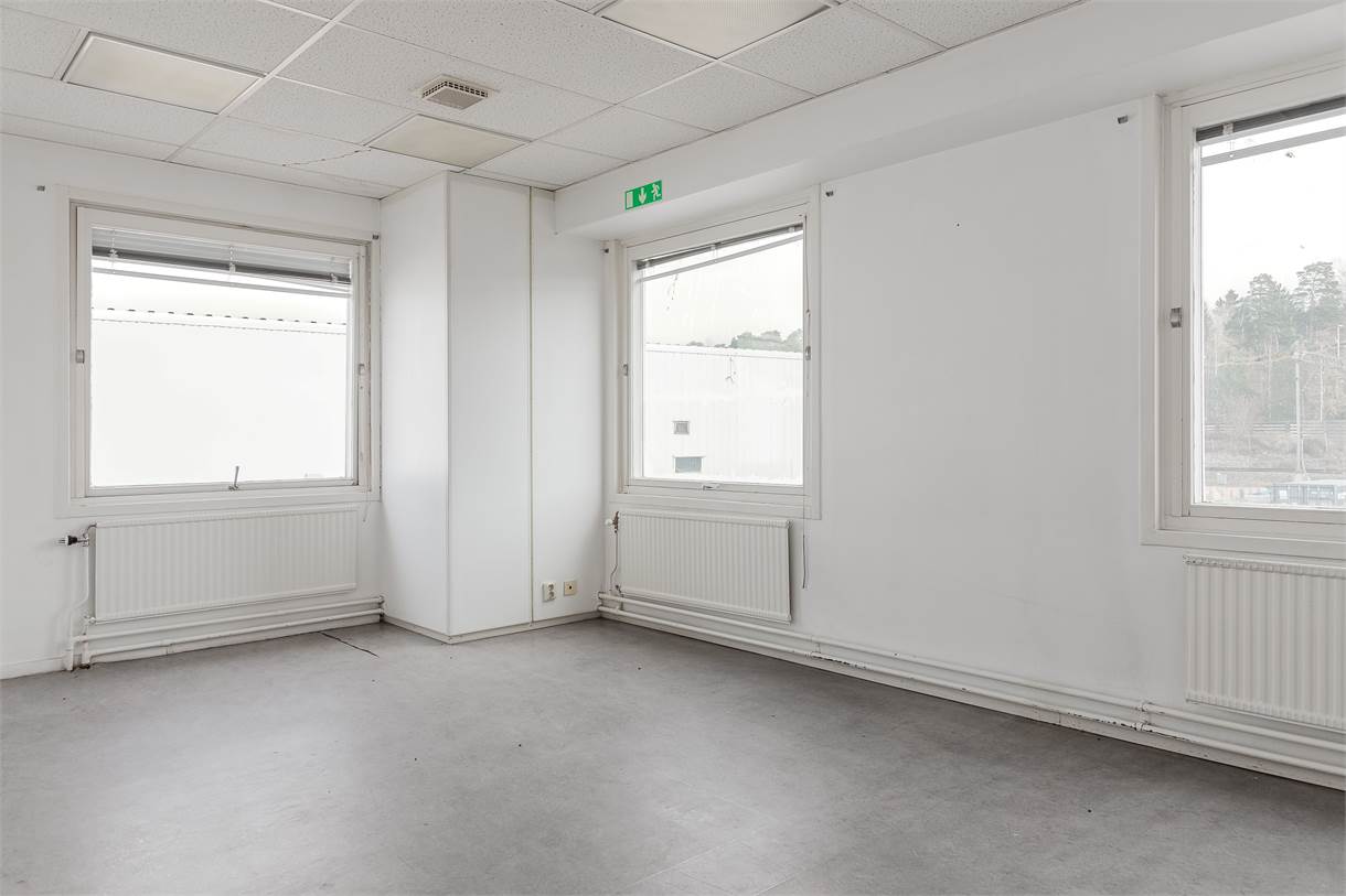 Tomt kontorsrum med vita väggar, tre fönster, radiatorer under varje fönster, grått golv och ett tak med infällda ljuspaneler.