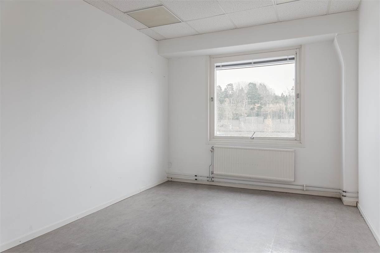 Tomt, omöblerat rum med vita väggar, grått golv, ett fönster med utsikt över träd och ett element under fönstret.