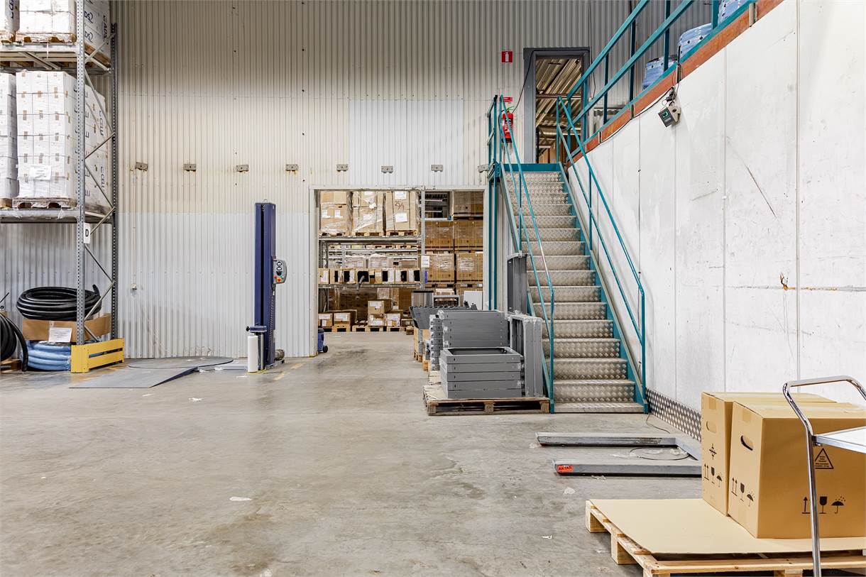 Lagerlokal med staplade lådor, metalltrappa, hyllor och en pallbock med en kartong på ett betonggolv.