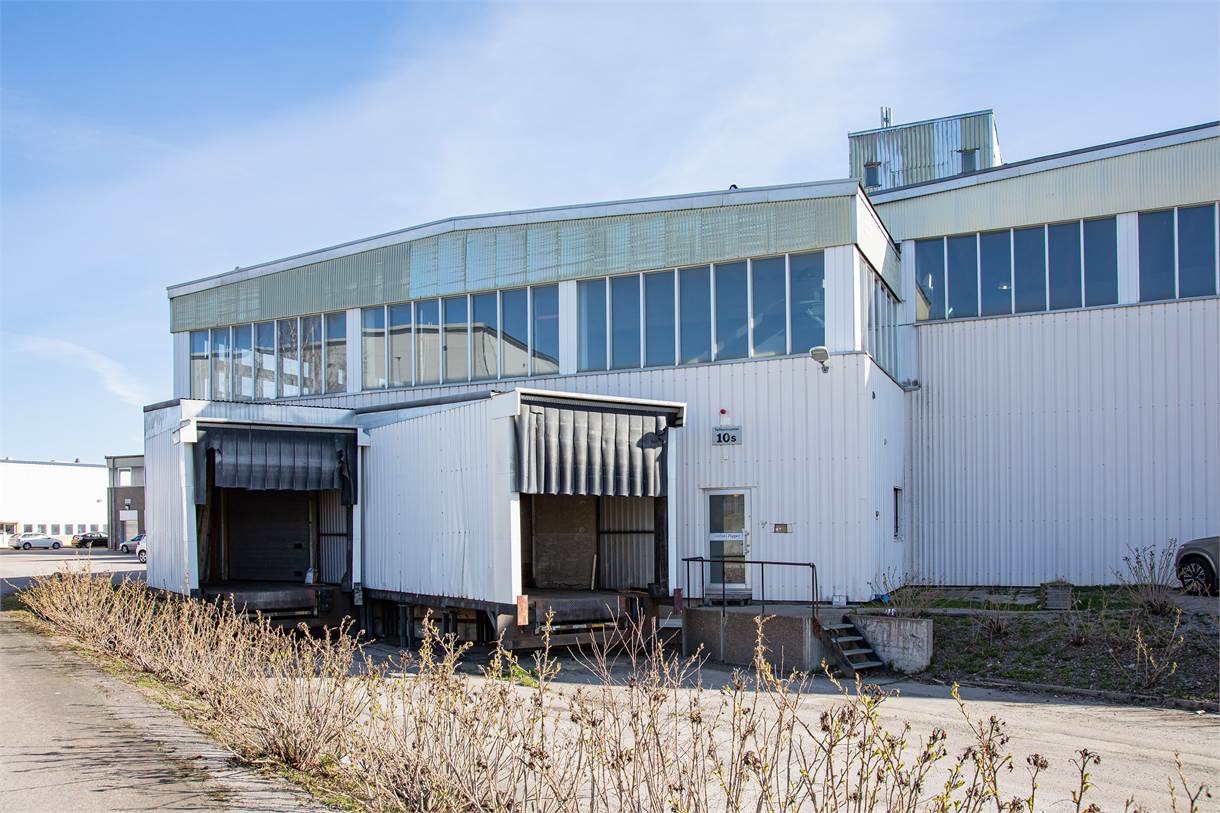 En vit industribyggnad med två lastkajer, metalltrappa och stora fönster under en klarblå himmel. Buskar kantar förgrunden.