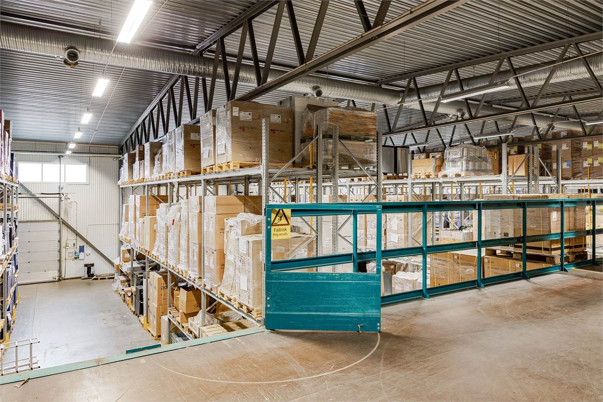 Interiörvy av en lagerlokal med hyllor fulla av lådor och pallar, metallbalkar i taket och en varningsskylt på ett tealfärgat räcke.