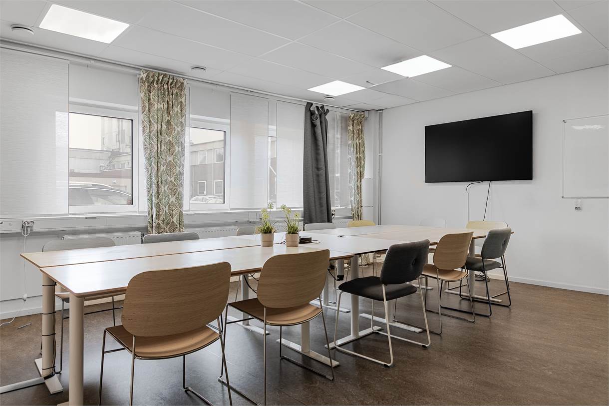 Ett modernt konferensrum med ett långt bord, olika stolar, krukväxter, en stor väggmonterad skärm och en whiteboardtavla. Naturligt ljus kommer in genom flera fönster.