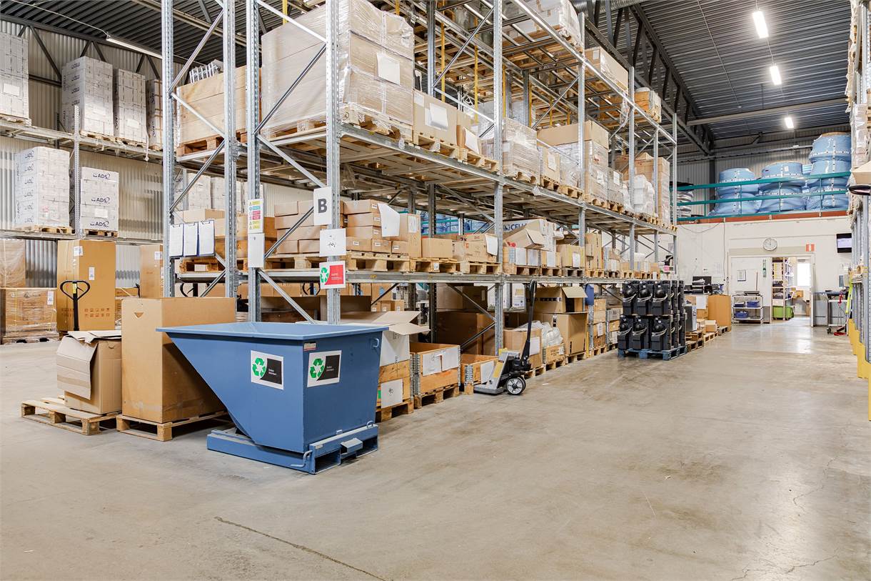 Lagerlokal med hyllor som rymmer olika lådor, en återvinningstunna i förgrunden och flera pallar och containrar staplade i hela utrymmet.