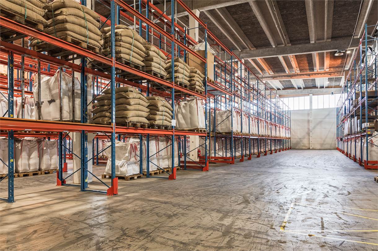 Lagerinredning med staplade pallar med stora påsar på metallhyllor, högt i tak och solljus som kommer in genom stora fönster och dörrar.