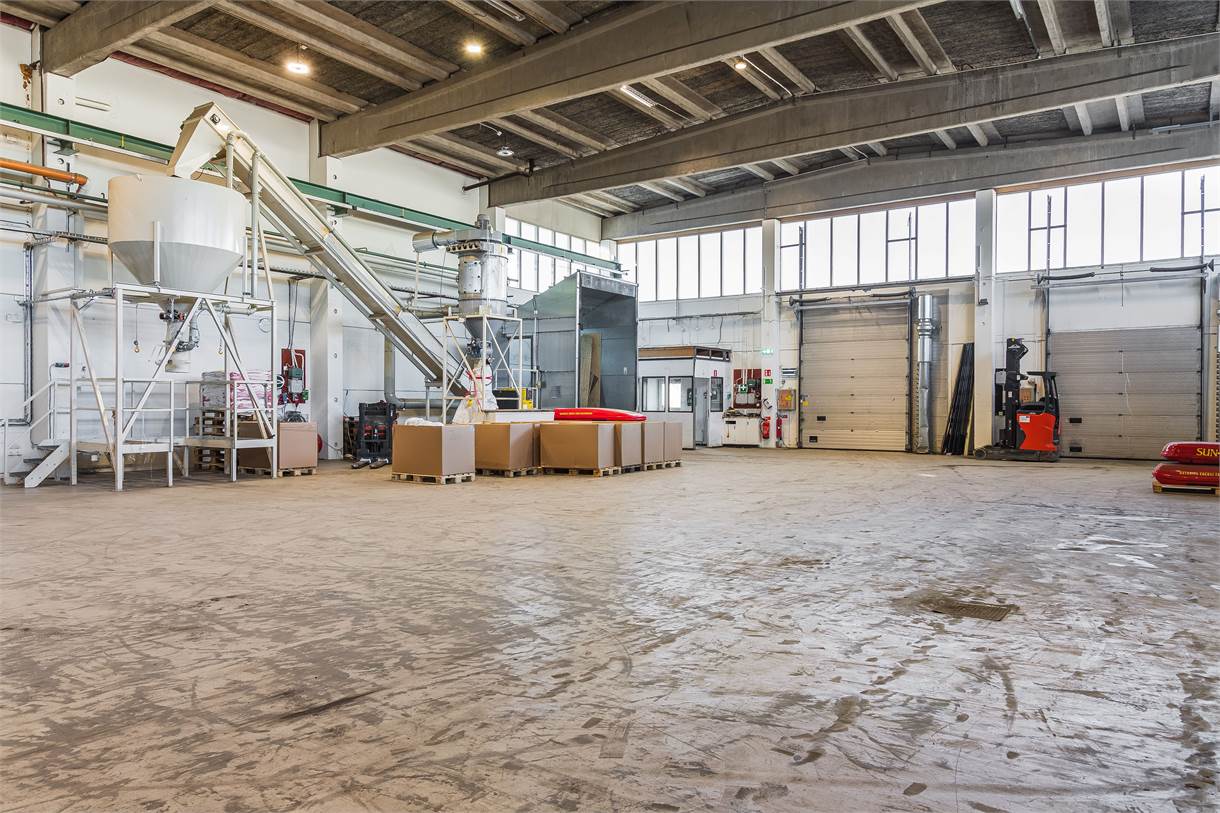 Rymlig industriell lagerlokal med maskiner, staplade lådor och palleterat gods på ett betonggolv under ett högt tak med synliga bjälkar.