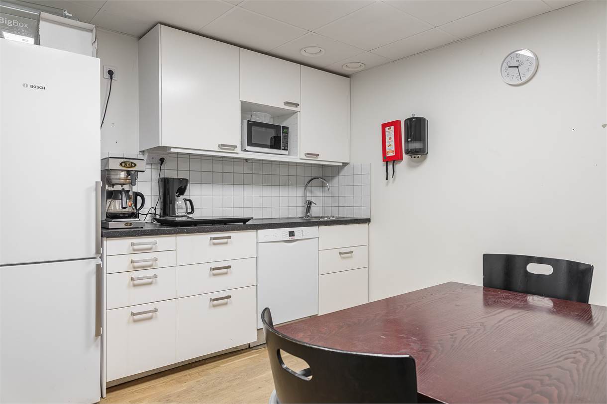 Litet kök med vita skåp, kylskåp, diskmaskin, kaffebryggare, mikrovågsugn, väggklocka, pappershandduksdispenser och ett mörkt träbord med svarta stolar.