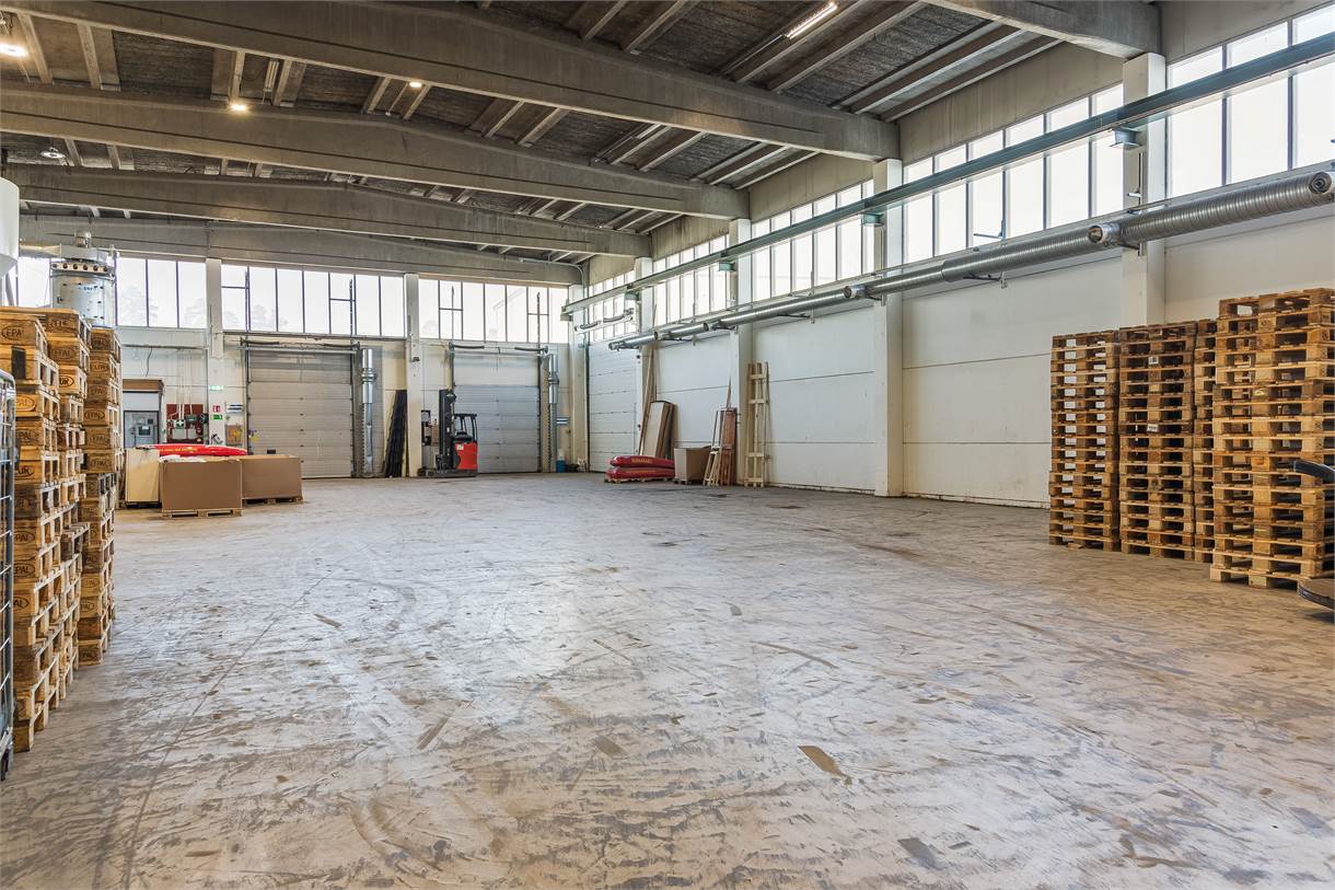 Rymlig lagerlokal med betonggolv, högt i tak, staplade träpallar och stora fönster som släpper in naturligt ljus.