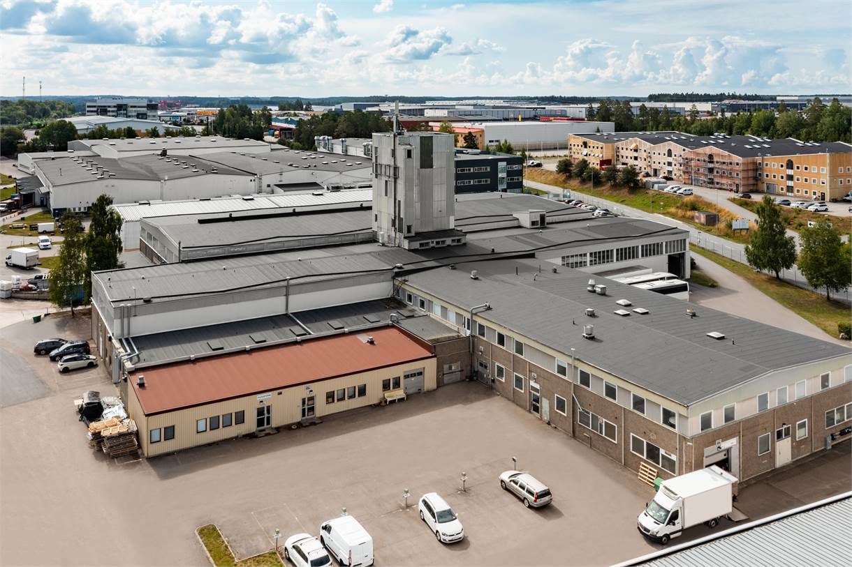Ett stort industriellt byggnadskomplex med flera byggnader med platta tak, omgivet av parkerade fordon och andra byggnader, under en delvis molnig himmel.