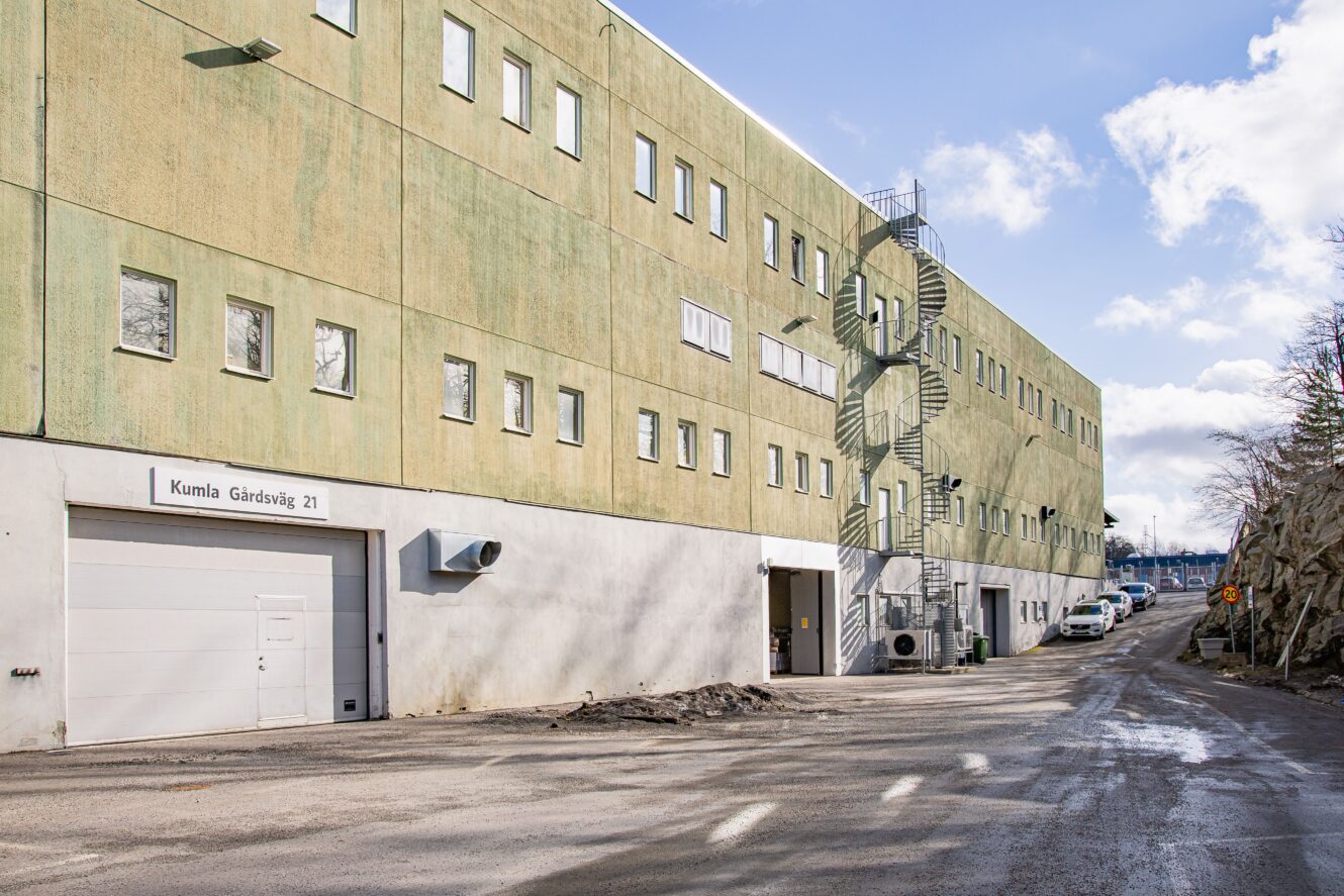 En stor ljusgrön industribyggnad med många fönster och en utvändig spiraltrappa; flera bilar uppradade längs den intilliggande vägen.
