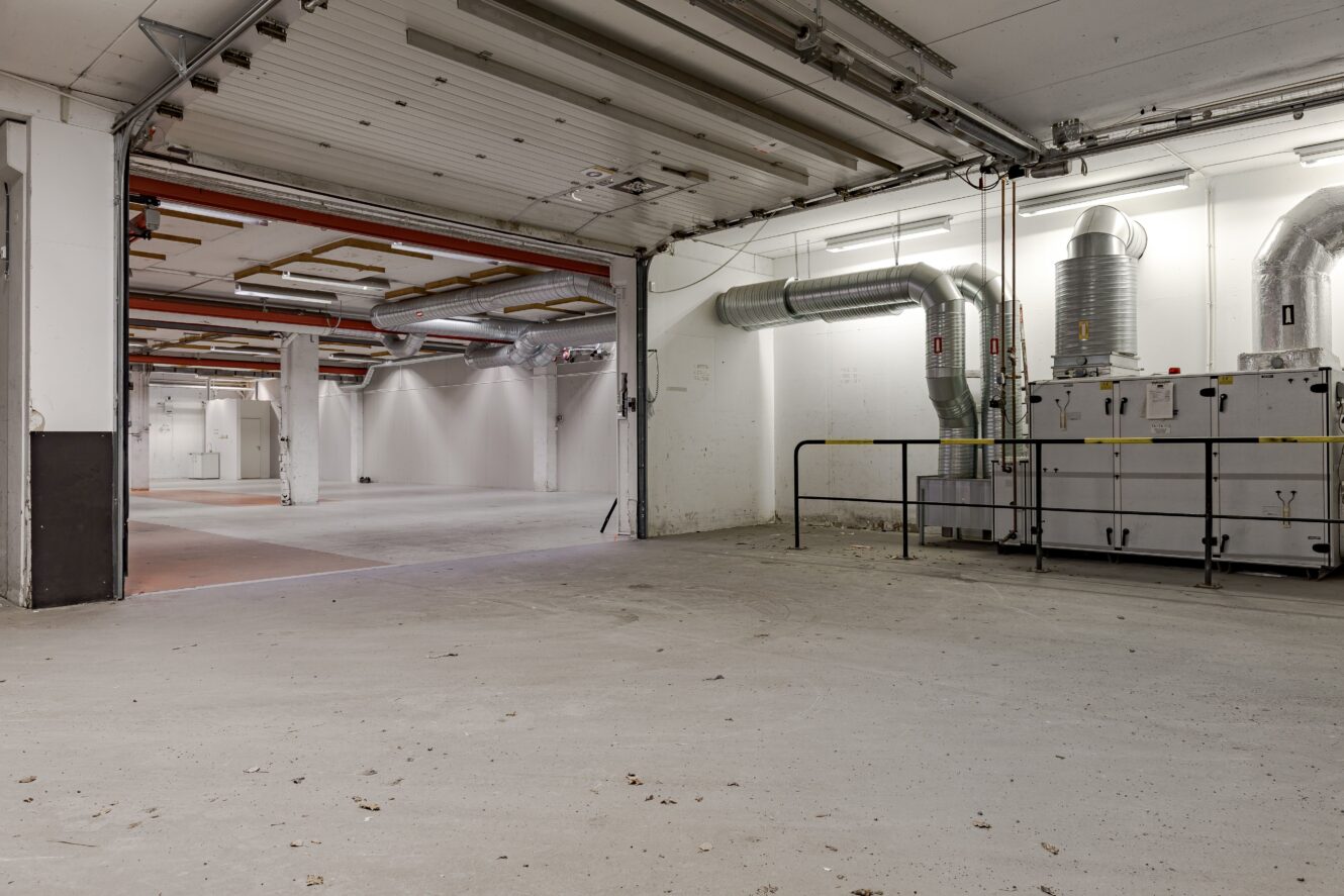 Stor, tom industrilokal med synliga kanaler, betonggolv, rörledningar, öppen garageport och varningstejp nära maskiner.