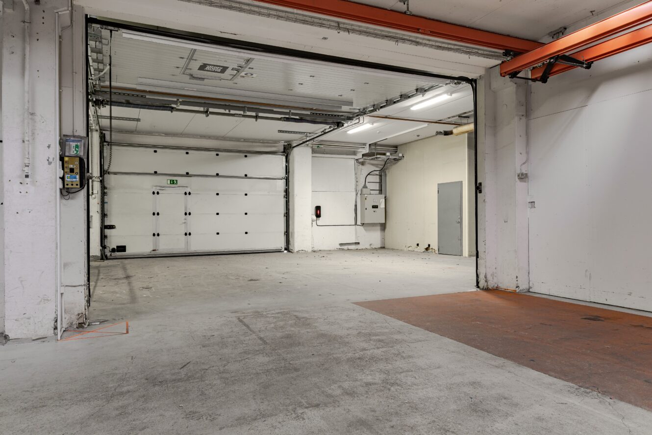 Tomt industrigarage med betonggolv, stor takskjutport, synliga rör, lysrörsbelysning och vita väggar med elpaneler.