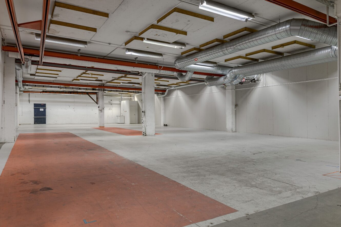 Stor tom industrilokal med exponerade kanalsystem, takbelysning, vita väggar, röda golvremsor och stödpelare överallt.