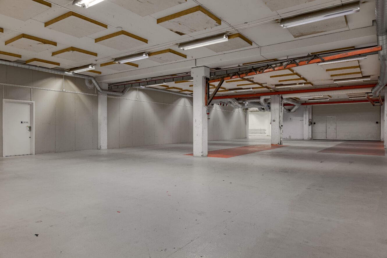 Stor, tom lagerlokal med vita väggar, synliga rör i taket, lysrör, stödpelare och flera stängda dörrar längs väggarna.