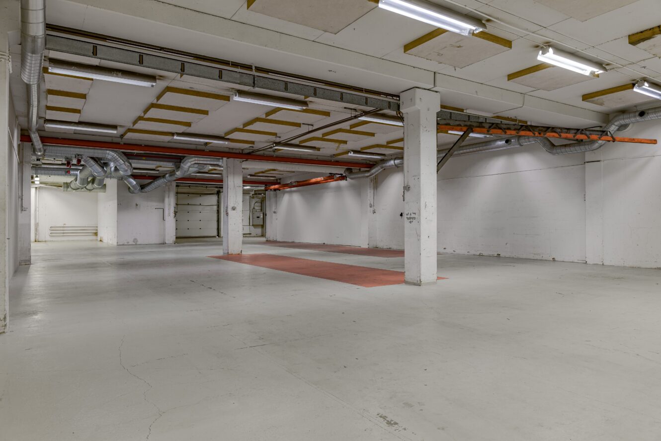 Rymlig, tom industrihall med vita väggar, synliga rör, lysrör och flera röda golvmarkeringar.