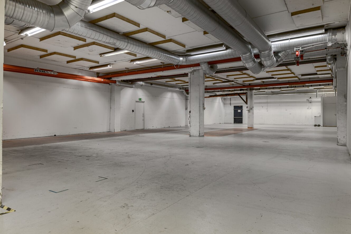 Stort tomt industrilager med synliga rör i taket, vita väggar, stödpelare, lysrör och betonggolv.