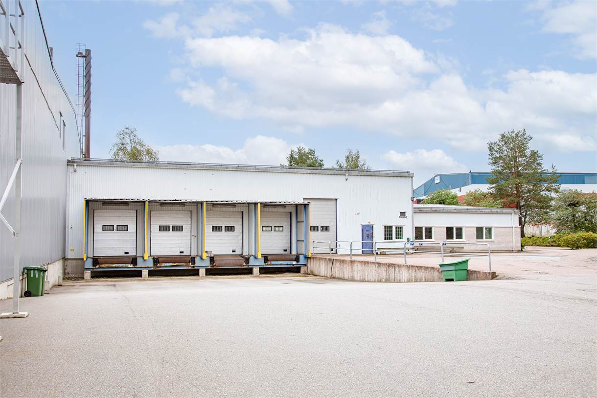 Ett lastkajsområde med fyra stängda garageportar, en betongramp och några träd och byggnader i bakgrunden under en delvis molnig himmel.