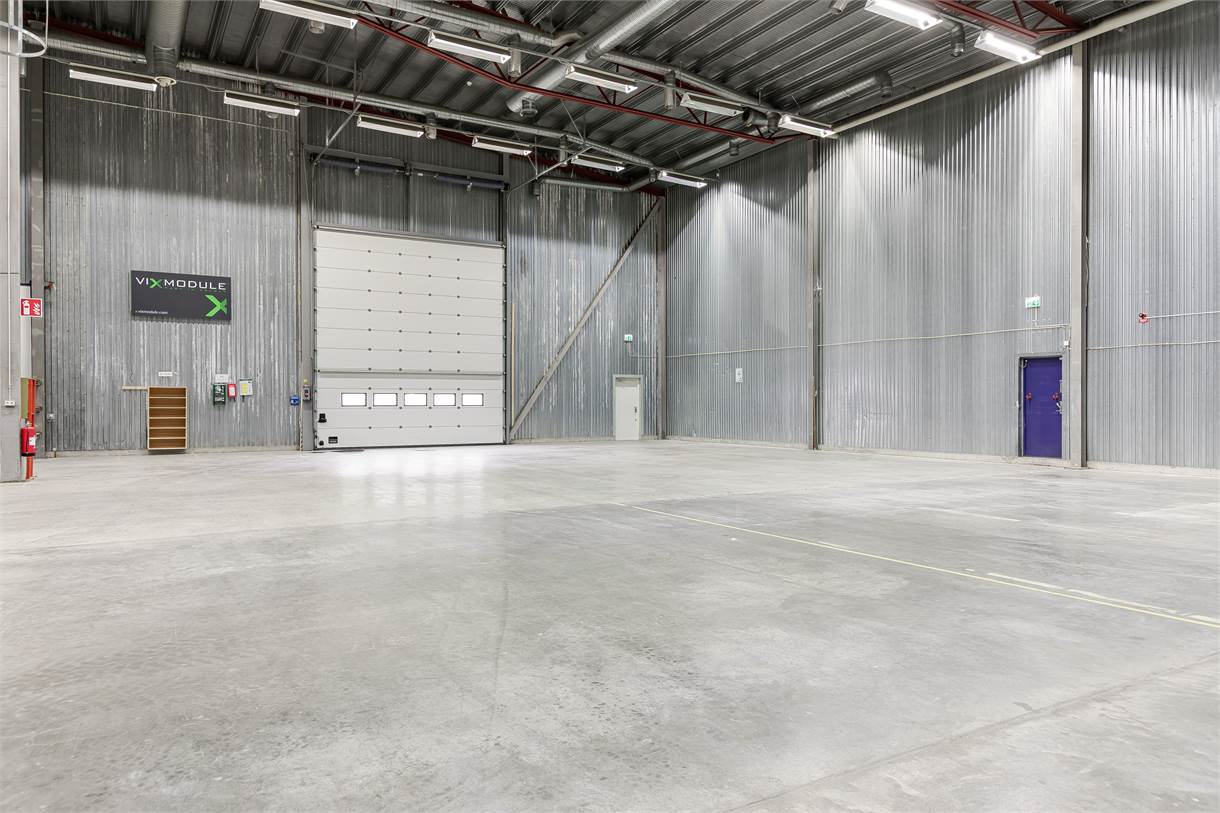 Interiör i en tom industrilokal med betonggolv, högt i tak, metallväggar, en stor garageport och lysrörsbelysning.
