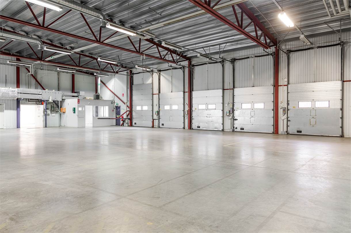 Rymlig, tom industrilagerinredning med betonggolv, metallväggar, högt i tak, synliga balkar och flera stora garageportar.