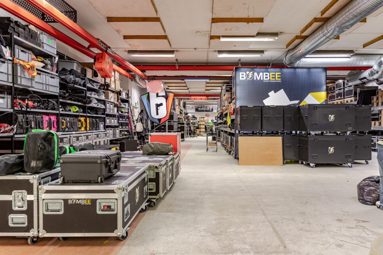 En rymlig lagerlokal med välorganiserade hyllor och stora utrustningslådor, varav flera märkta BOMBEE, under starka taklampor.