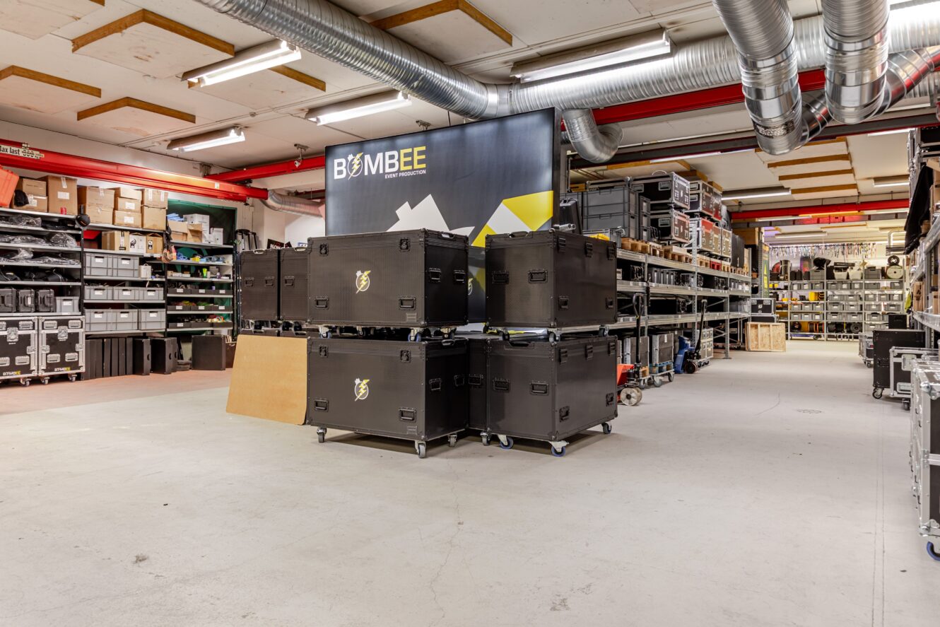 Ett förråd med stora svarta utrustningslådor på hjul, metallhyllor med olika lådor och utrustning samt en banderoll märkt Bombee.