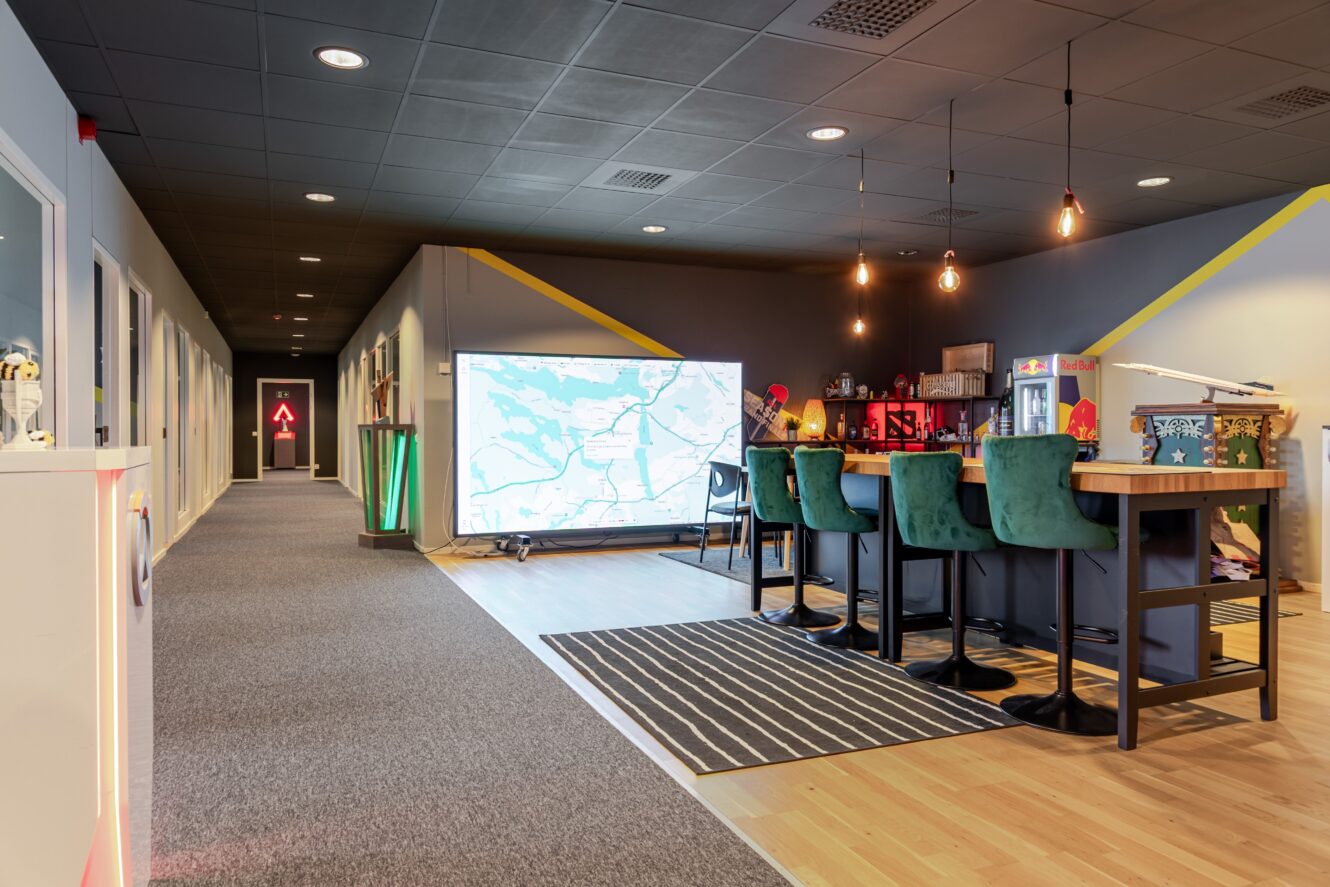 Moderna kontorslokaler med höga bord och gröna stolar, en stor väggskärm med en karta, hängande lampor och en intilliggande hall med kontorsdörrar.