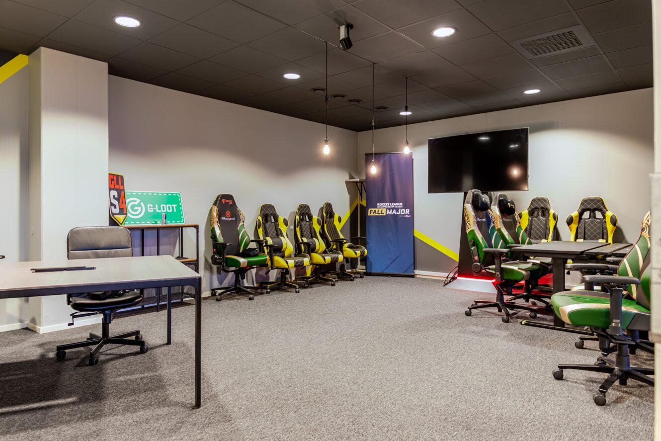Ett modernt gamingrum med flera gamingstolar, skrivbord, en stor väggmonterad TV, hängande lampor och esportrelaterade skyltar.