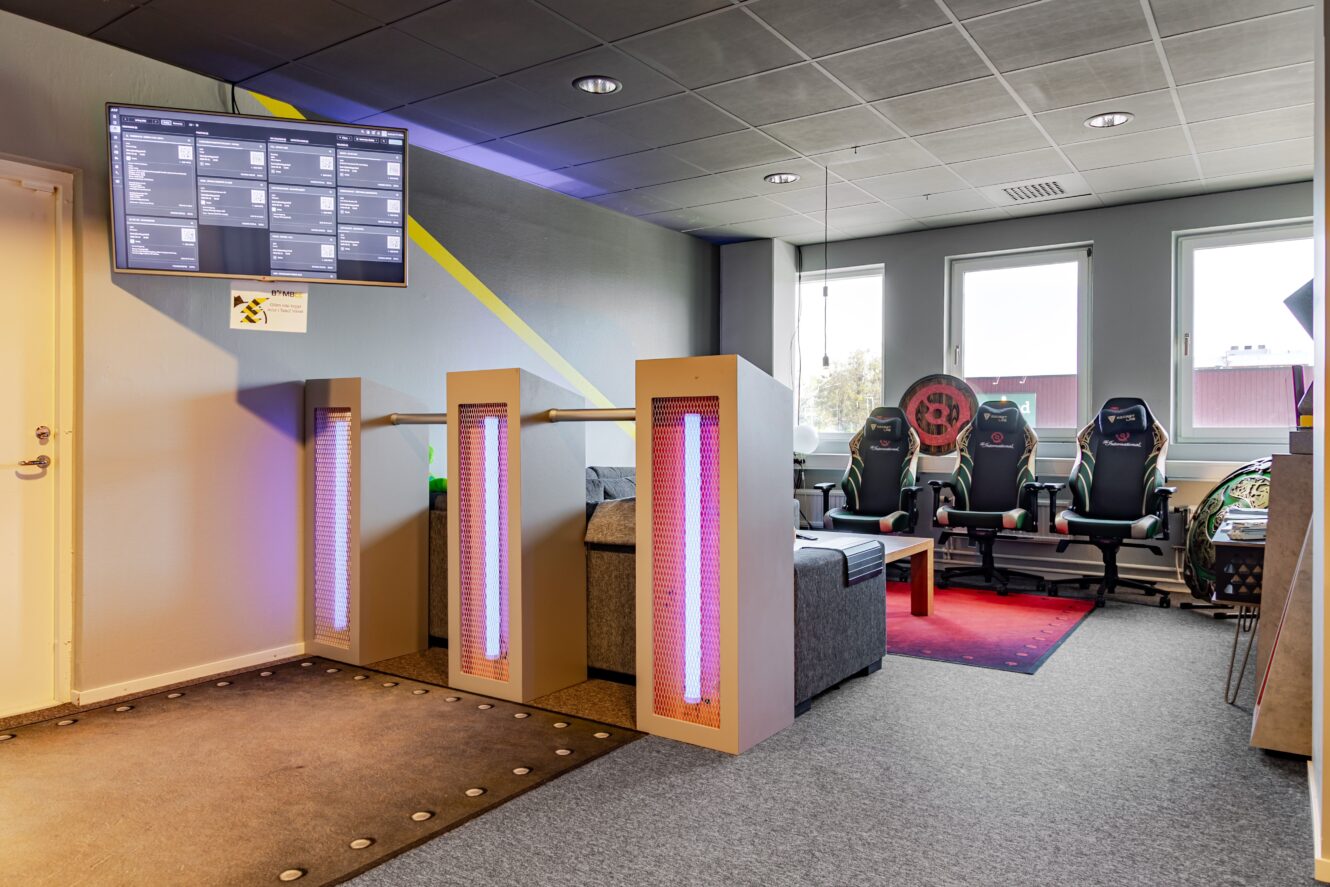 En modern gaminglounge med tre gamingstolar, en röd matta, två upplysta skiljeväggar, en soffa och en väggmonterad skärm som visar scheman nära ett fönster.