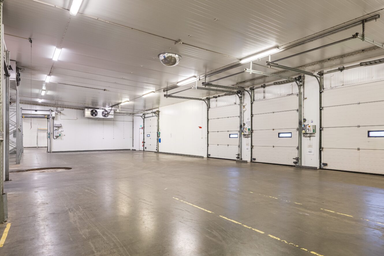 Stort, tomt industrilager med flera takskjutportar, vita väggar, högt i tak och lysrörsbelysning.