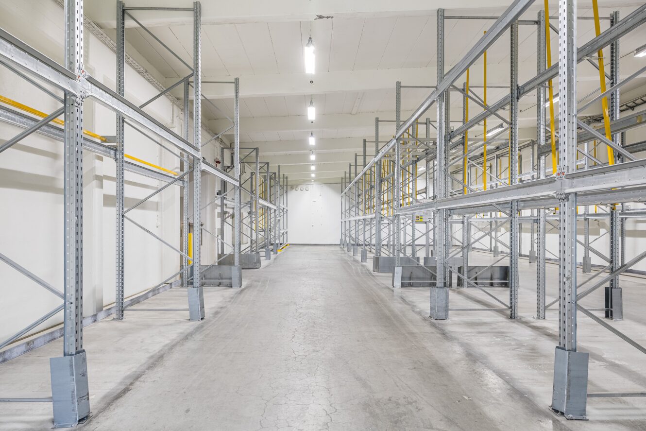 Tom lagerlokal med metallhyllor, betonggolv och lysrörsbelysning i en ren och organiserad miljö.