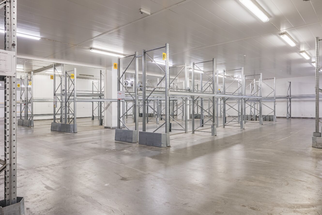 Tomt industrilager med lagerhyllor i metall, betonggolv och stark takbelysning.