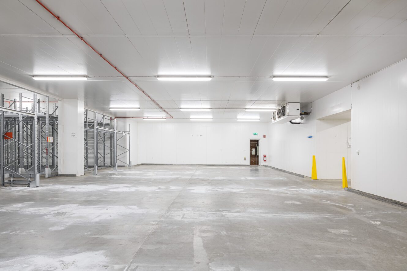 Tomt industriellt förråd med betonggolv, väggar med vit panel, metallhyllor till vänster, lysrör i taket och två gula pollare nära den bakre väggen.