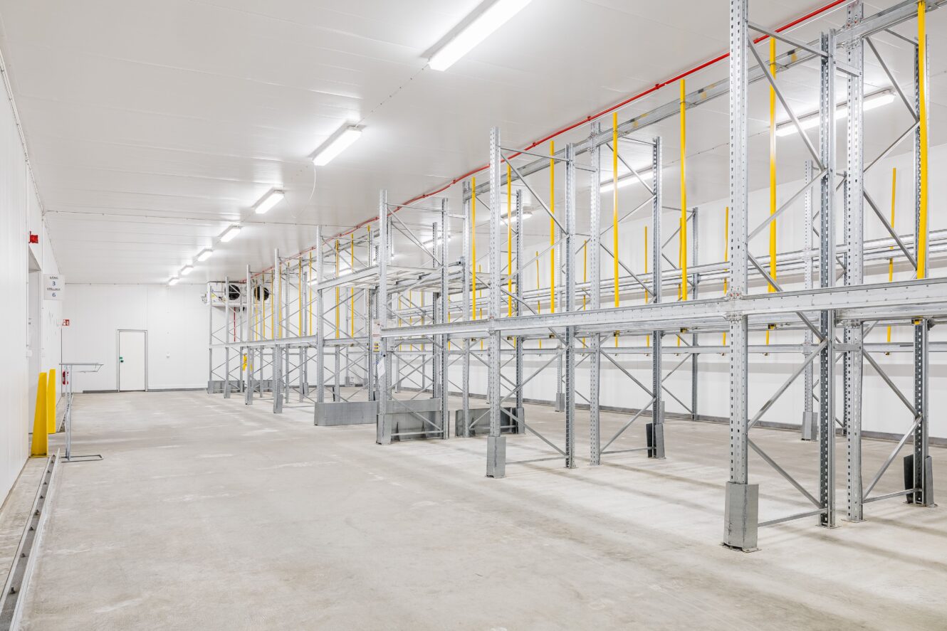 Tomt industrilager med lagerhyllor i metall, betonggolv och stark takbelysning.